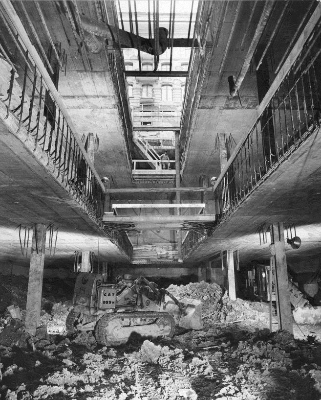 Der Blick der Schwarz-weiss-Fotografie ist von einem der Untergeschosse nach oben gerichtet, wobei die in der Mitte noch geöffneten Decken die Sicht auf die Fassade des oberirdischen Bibliotheksgebäudes zulassen. Die Arbeiten befinden sich im Rohbau: Auf der Ebene der Kamera schaufelt ein Bagger Erdhaufen, und überall sind aus den Betondecken ragende Armierungseisen zu erkennen.